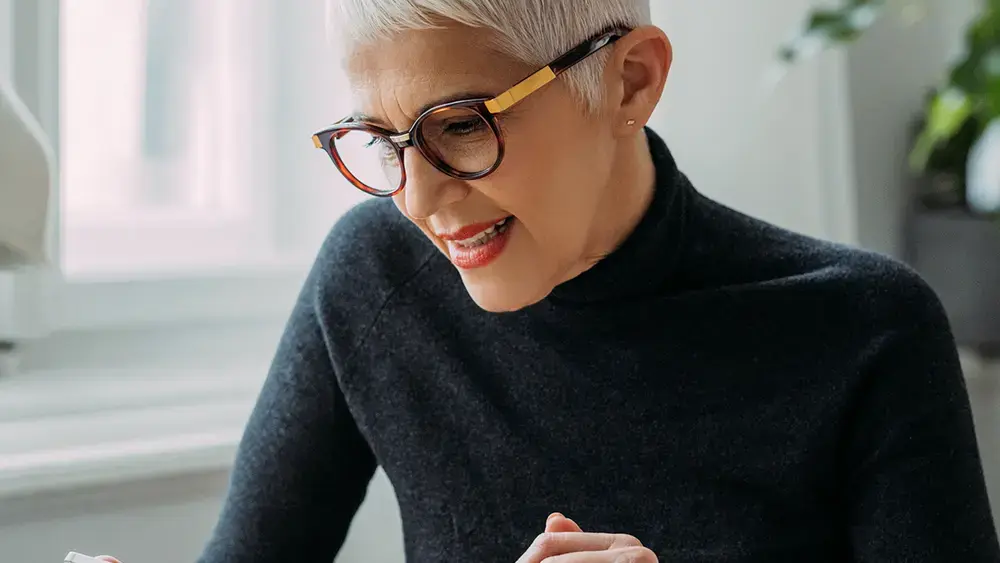 Une femme porte des lunettes et un col roulé noir, et utilise une tablette à écran tactile.