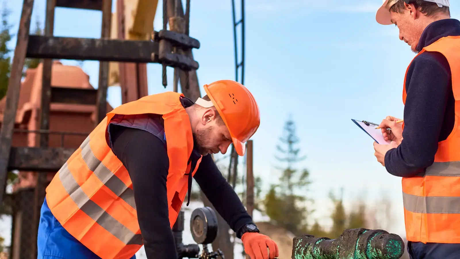 Deux travailleurs en gilet orange inspectent des équipements près d'une plateforme pétrolière.